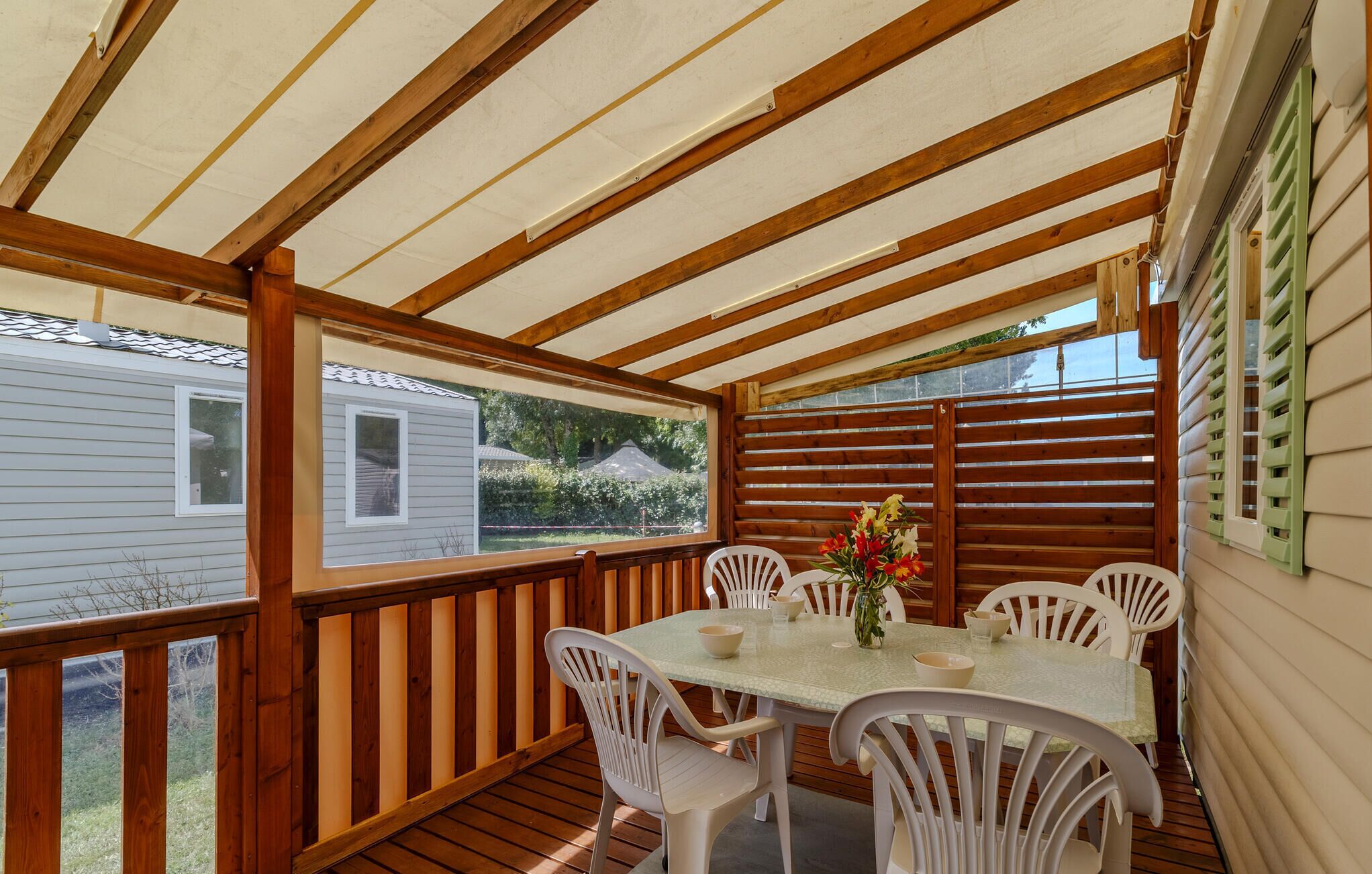 En overdækket terrasse med et bord og stole, træbjælker og udsigt til et hus.