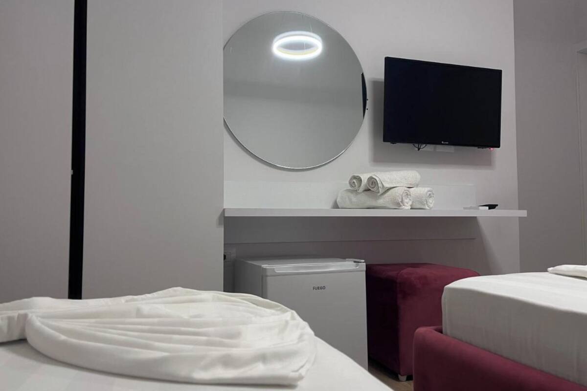 Une chambre d’hôtel avec un lit, un petit minibar, un téléviseur fixé au mur, un miroir et une serviette sur le comptoir.
