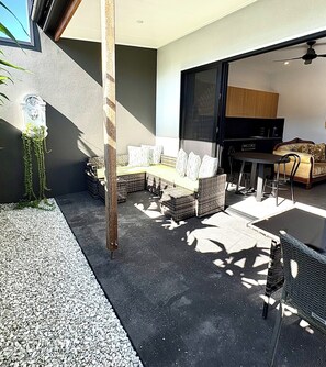 Un espace extérieur aménagé en terrasse, avec des meubles en osier, une table à manger et un coin cuisine visible à travers une porte ouverte.