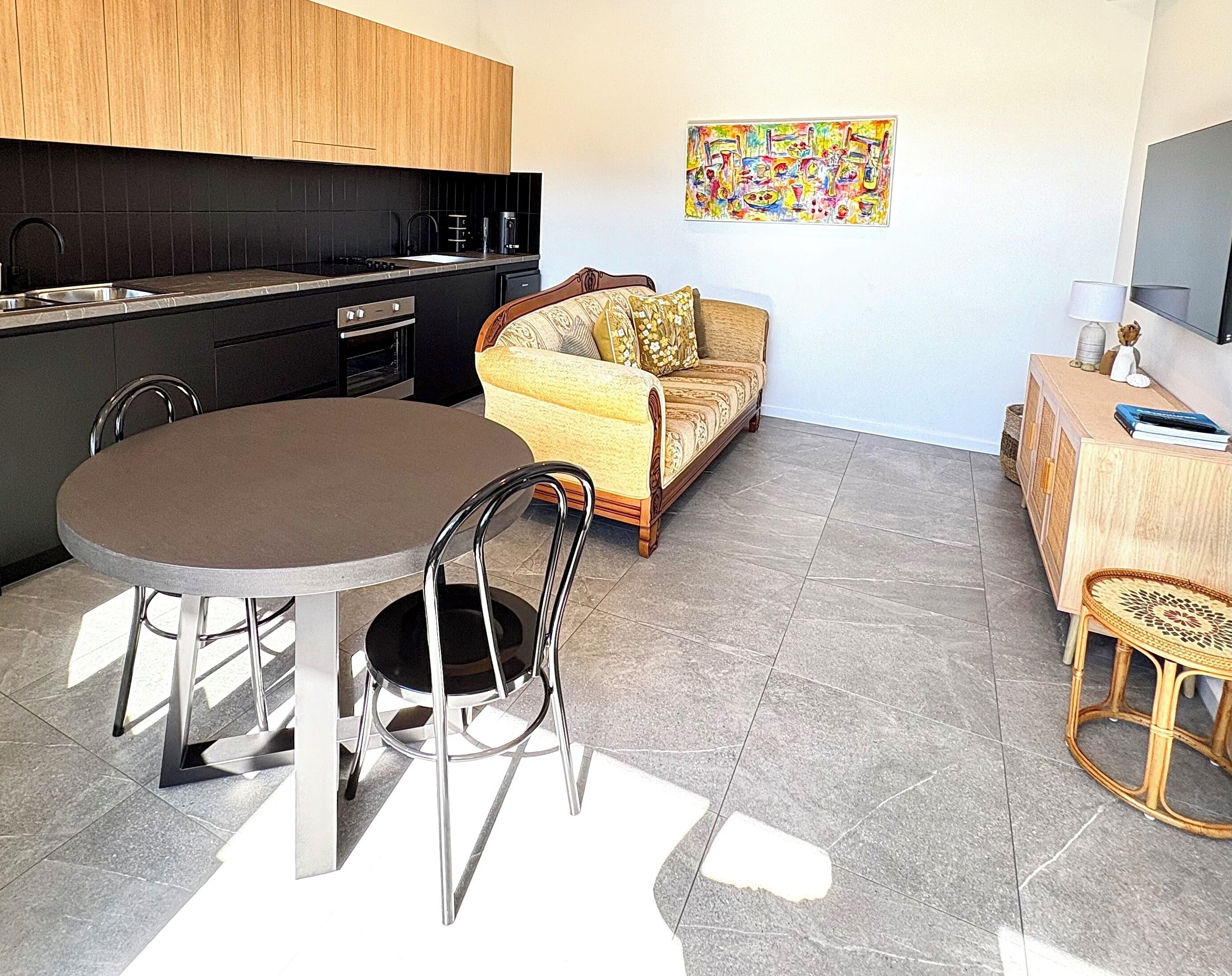 Une cuisine compacte dotée d’appareils électroménagers modernes, un coin repas avec une table ronde et des chaises, un canapé et une petite table d’appoint en bois avec une lampe.