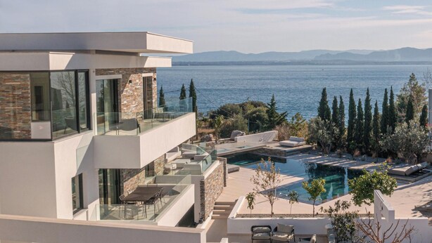 A modern multi-story building with a glass balcony overlooking a pool and a body of water. - Elia Cove Luxury Villas & Residences (Loutraki-Agioi Theodoroi)