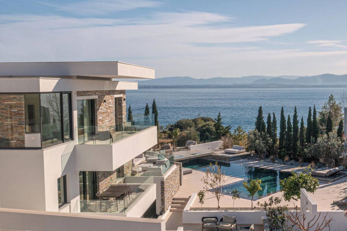 A modern multi-story building with a glass balcony overlooking a pool and a body of water.