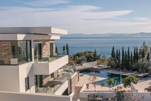 A modern multi-story building with a glass balcony overlooking a pool and a body of water.