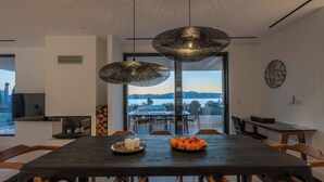 A modern dining area with a dark wooden table, six wooden chairs, two hanging pendant lights, and a view of a lake and mountains through large glass doors. - Elia Cove Luxury Villas & Residences (Loutraki-Agioi Theodoroi)