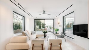 A modern living room with a sofa set, two armchairs, a coffee table, and a TV. Large windows offer views of a garden and a cityscape. - Elia Cove Luxury Villas & Residences (Loutraki-Agioi Theodoroi)