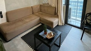 A modern living room with a beige sectional sofa, a black coffee table with decorative items, a cityscape painting on the wall, and a view of a building through a large window.