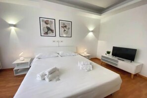 A modern hotel room with a large bed, white bedding, two wall-mounted light fixtures, a flat-screen TV, and a small plant on a white console table. - Spacious and Elegant Apartment on Viale Libia
 (Roma)