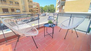 En balkon med to hvide stole af metaltråd, et lille rundt bord med en potteplante, og udsigt over en byvej med parkerede biler og bygninger.