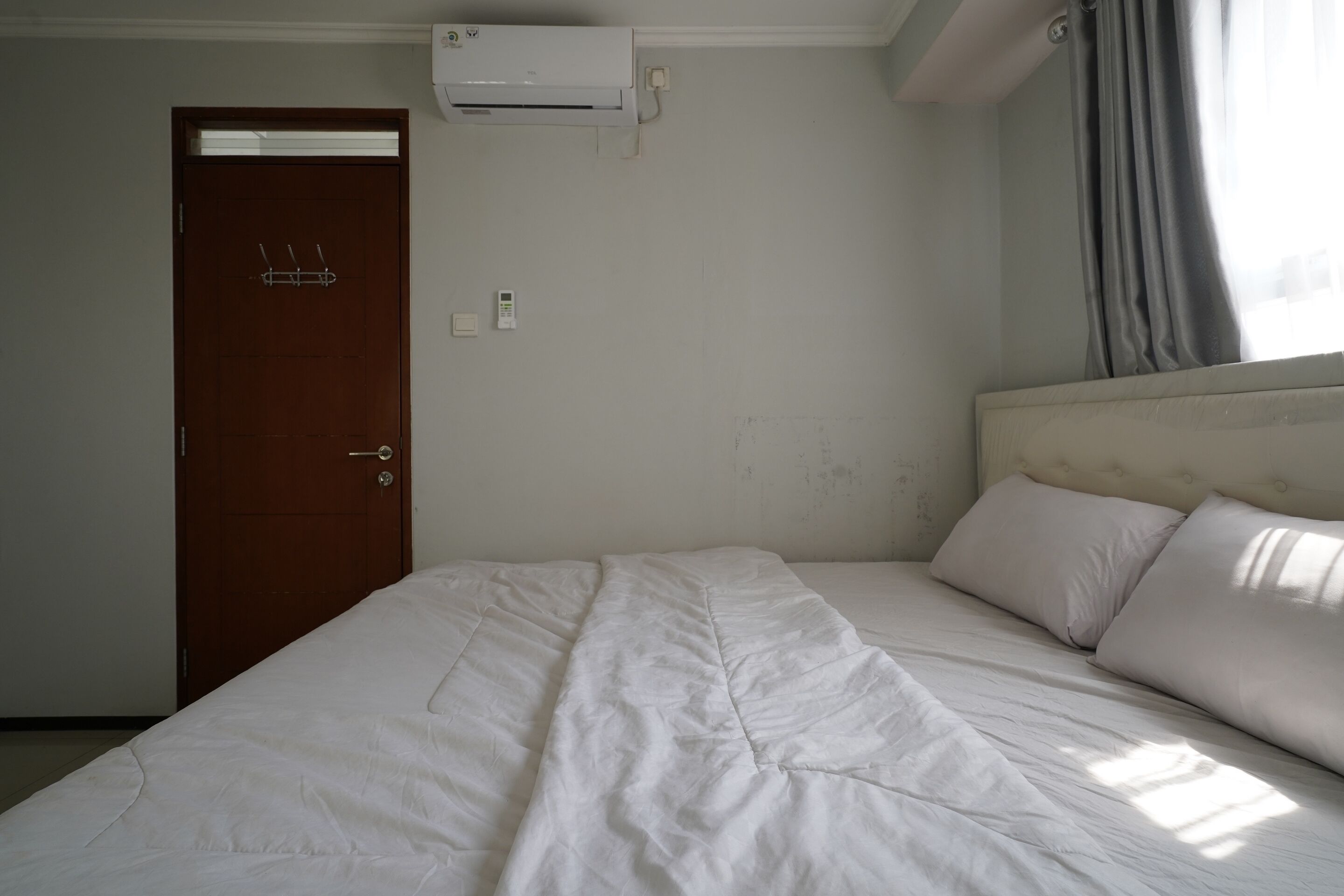 Una habitación con cama, puerta, equipo de aire acondicionado y ventana con cortinas.