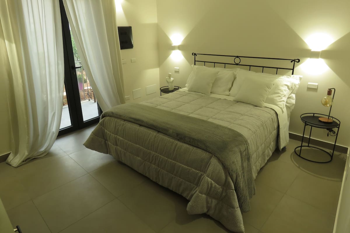 Quarto com cama, mesa de cabeceira, luminária e vista do exterior através de uma janela com cortinas.