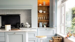 Une cuisine avec des meubles de couleur bleu clair, un parquet en bois disposé en chevrons, un tabouret rond blanc et une fenêtre donnant sur un paysage verdoyant.