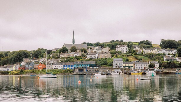 Une ville côtière avec des maisons le long du littoral, une église dont le clocher est particulièrement visible, et des bateaux amarrés au quai.