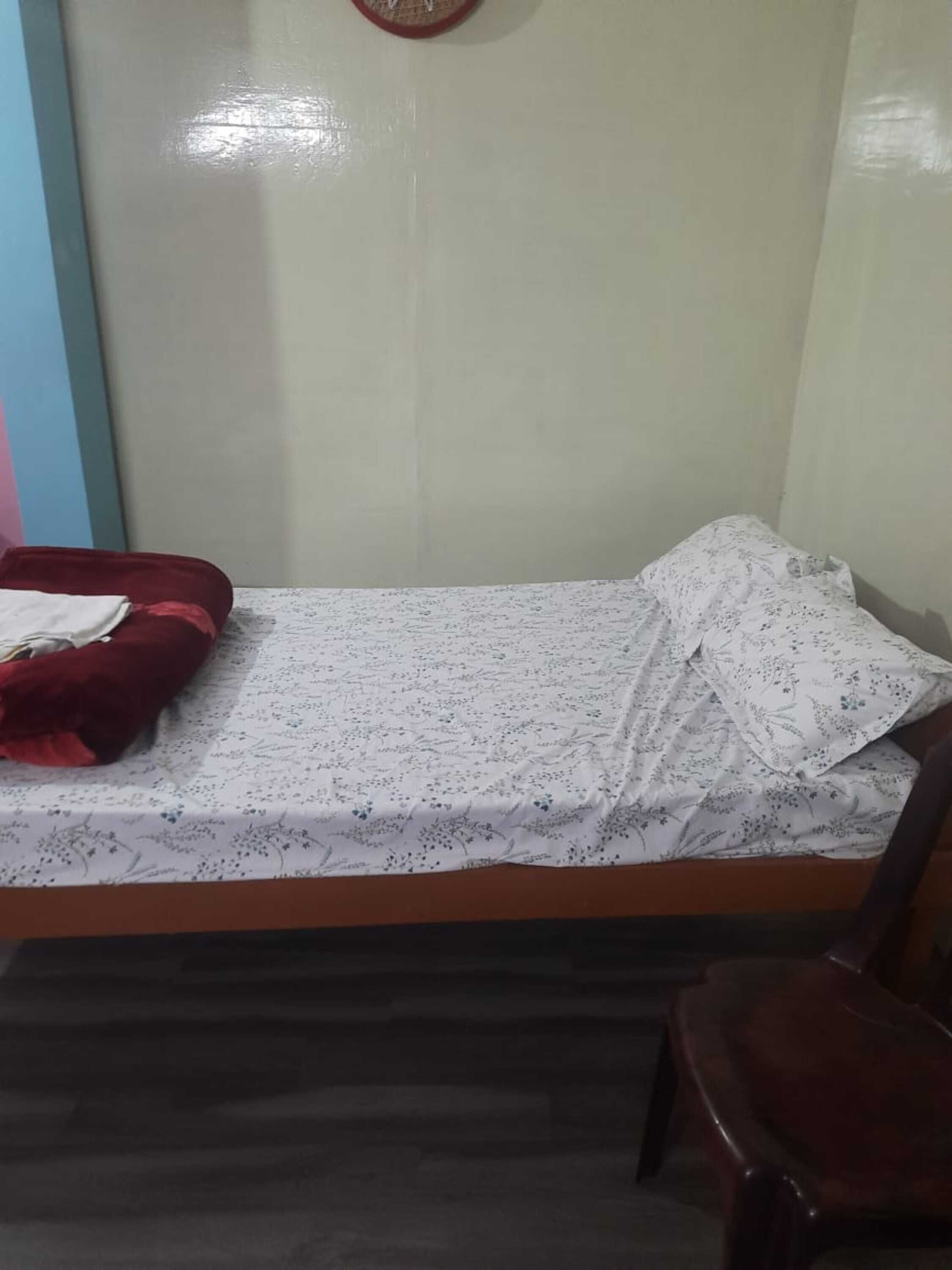 Une petite chambre avec un lit simple, un cadre en bois, une couvre-lit à motifs floraux et un oreiller rouge.