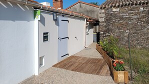 A narrow alleyway with a white building on one side and a stone wall on the other. There is a wooden deck leading to the building, and a small garden with flowers.