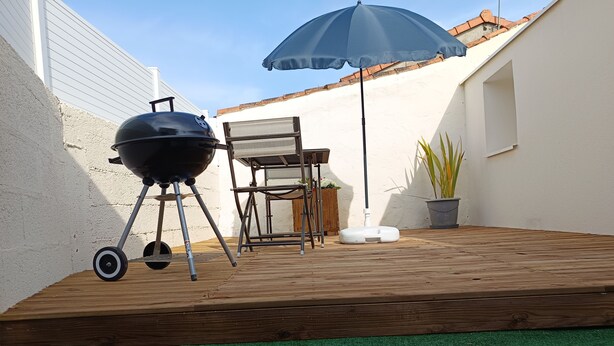 A patio with a grill, a chair, a table, and a potted plant.