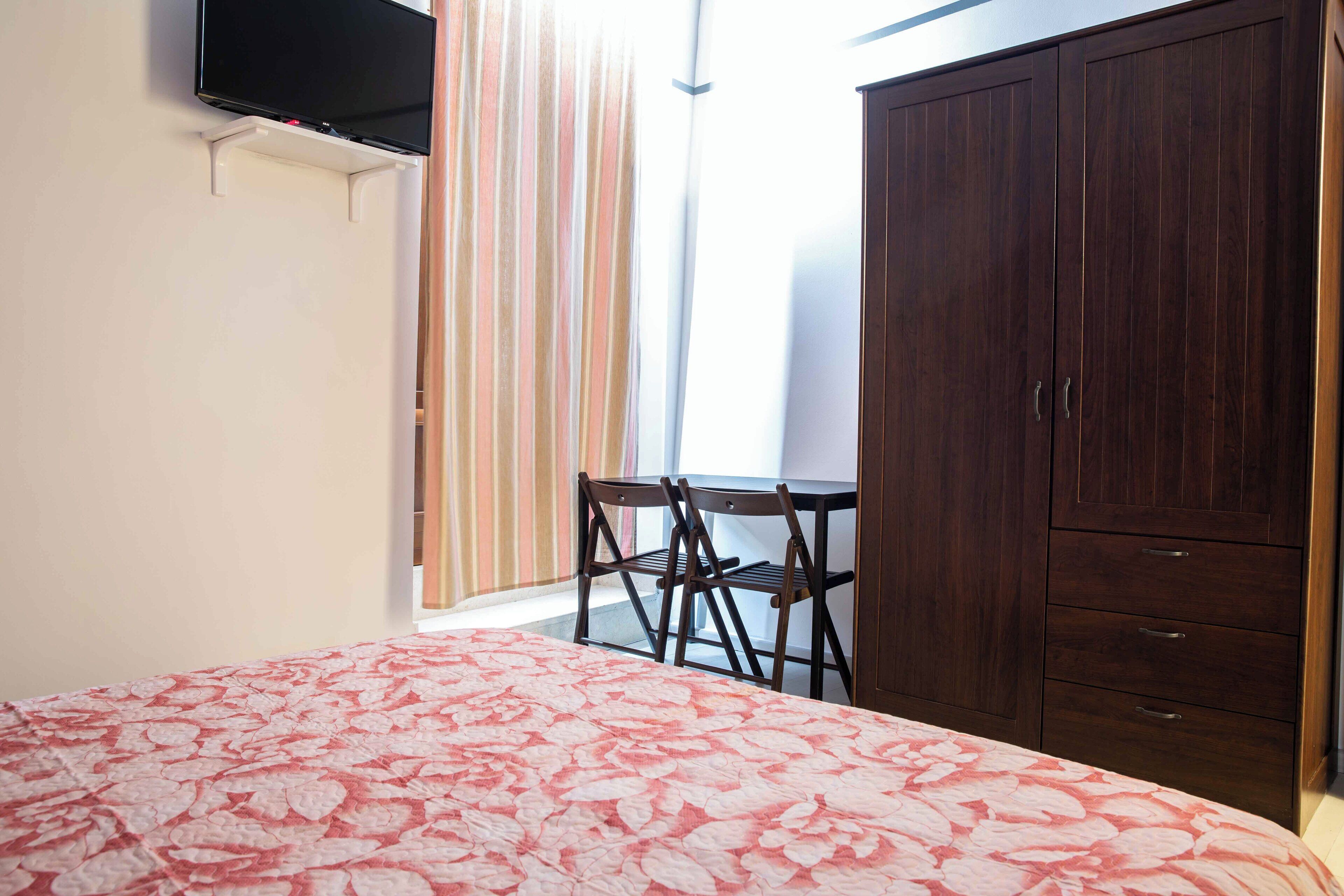 A bedroom with a bed, a wardrobe, a TV mounted on the wall, and a window with curtains.