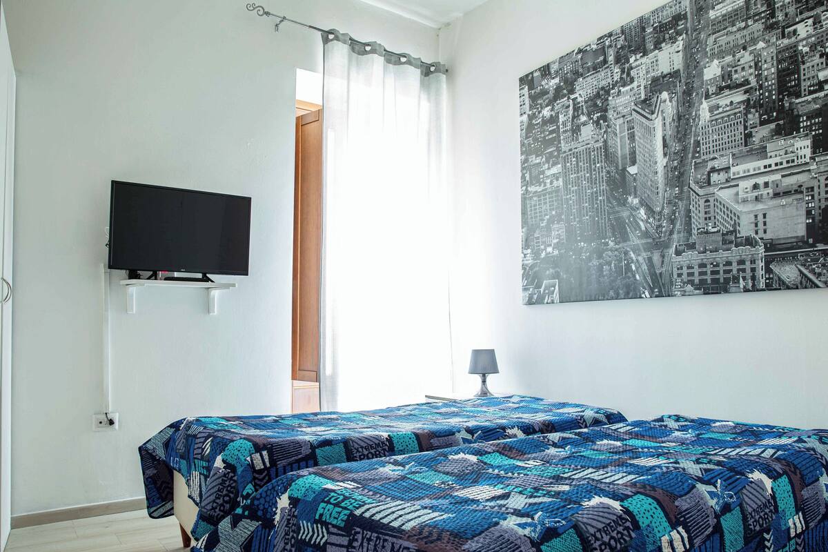 Un dormitorio con cama, televisor, un cuadro de paisaje urbano y una ventana con cortinas.