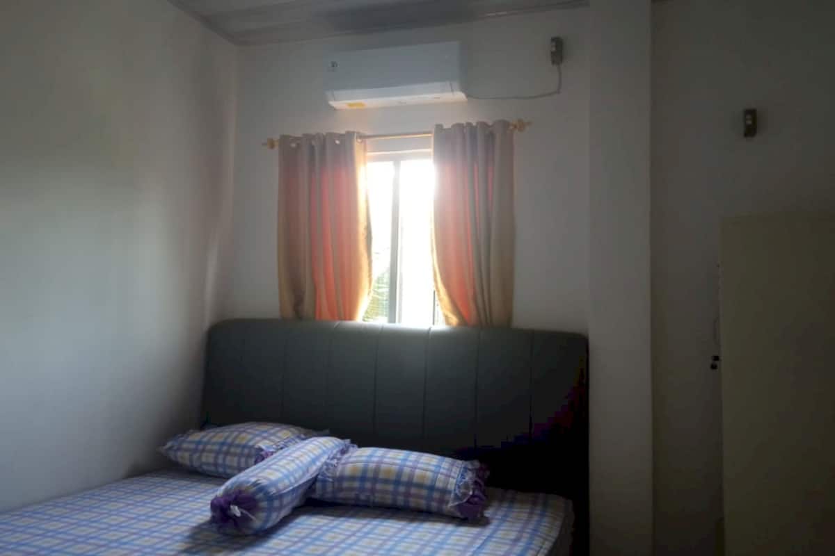 A small, single-bed room with a window, curtains, and an air conditioning unit.
