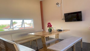 Een kamer met een houten tafel en bankjes, een aan de muur bevestigde televisie en een raam met uitzicht op gebouwen.