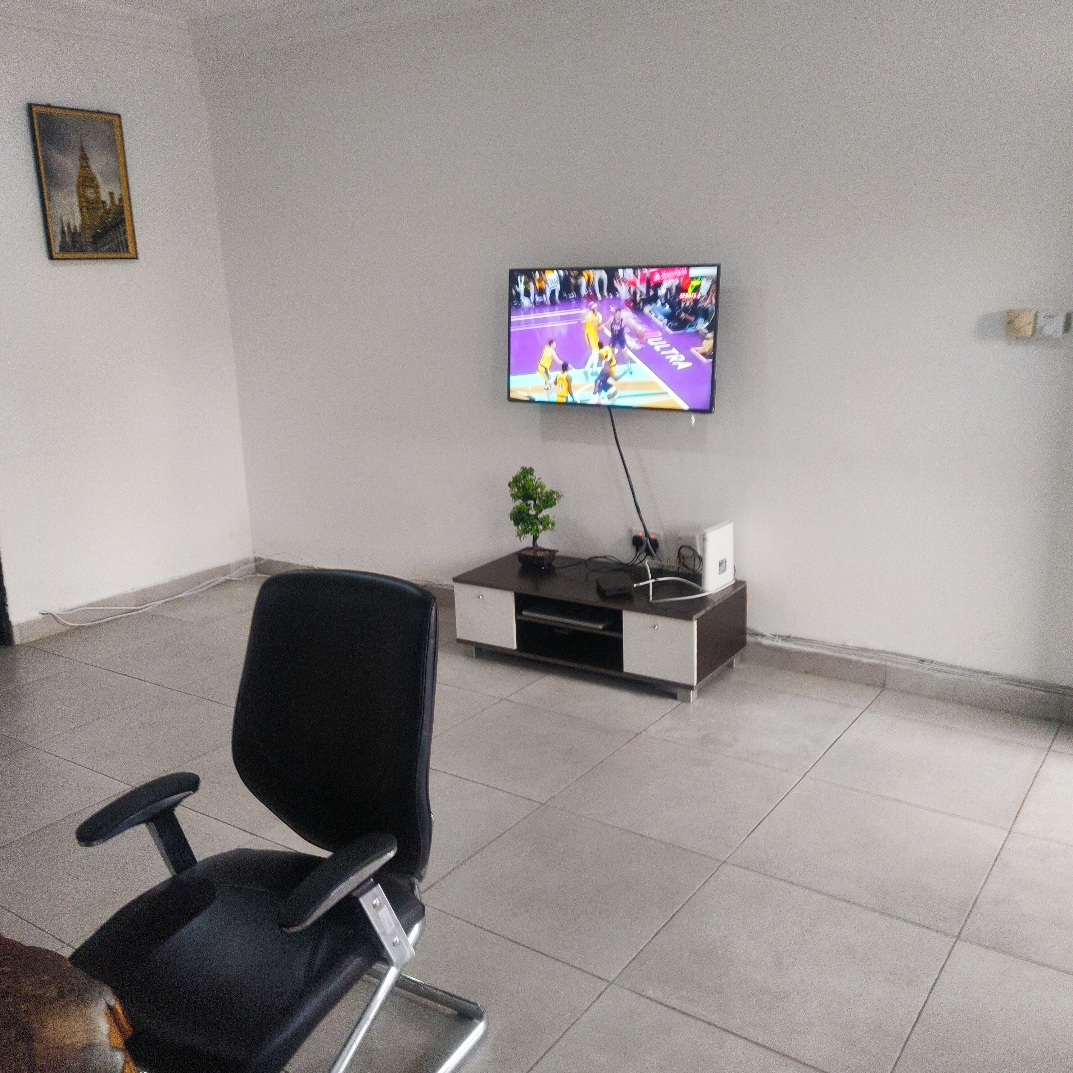 A modern office space with a flat-screen TV mounted on the wall, a black office chair, a small plant on a side table, and a framed picture of a building.