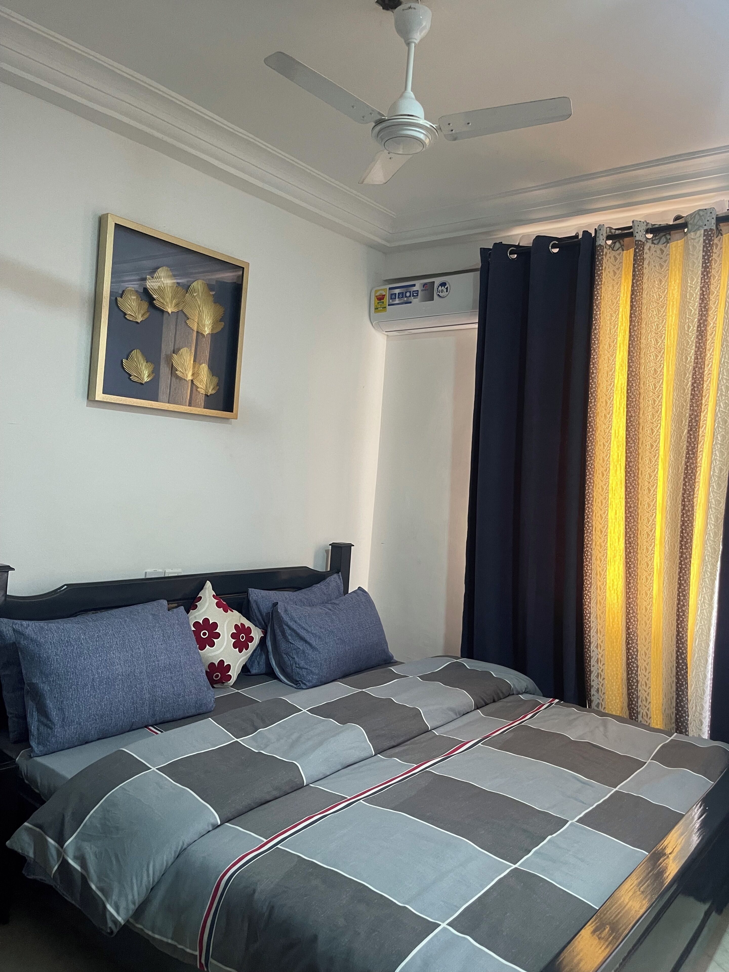 A bedroom with a bed, a ceiling fan, a framed artwork, a wall-mounted air conditioner, and patterned curtains.