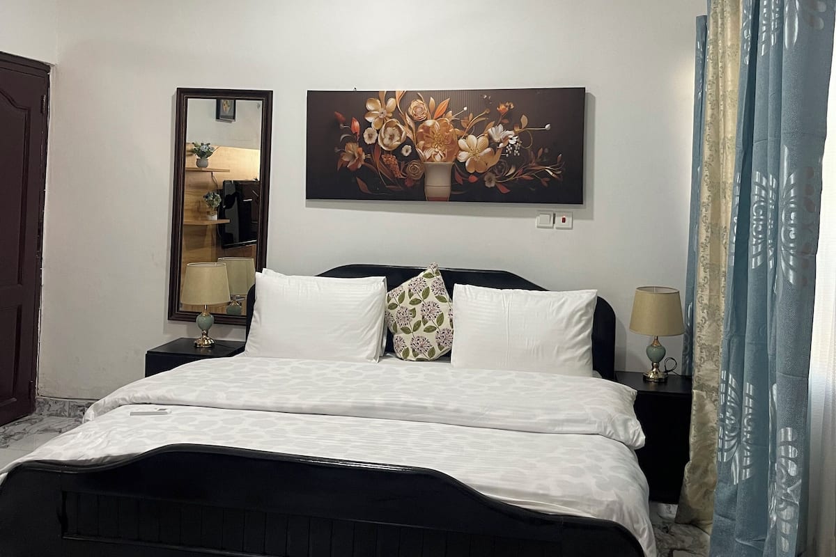 Une chambre d’hôtel comprenant un lit, un ventilateur de plafond, un climatiseur mural, un miroir et une œuvre d’art à motifs floraux.