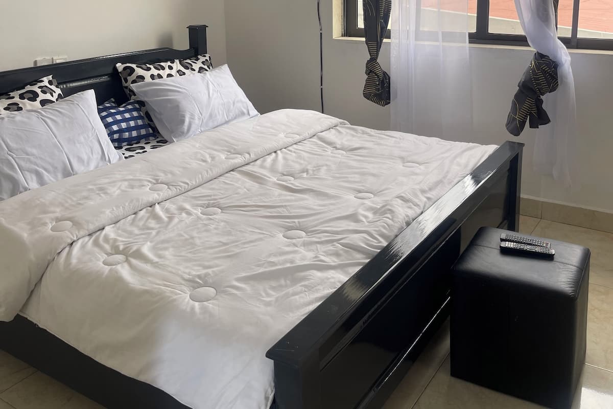 Un lit bien fait, avec un cadre noir et du linge de lit blanc, un petit pouf noir et un ventilateur de plafond.