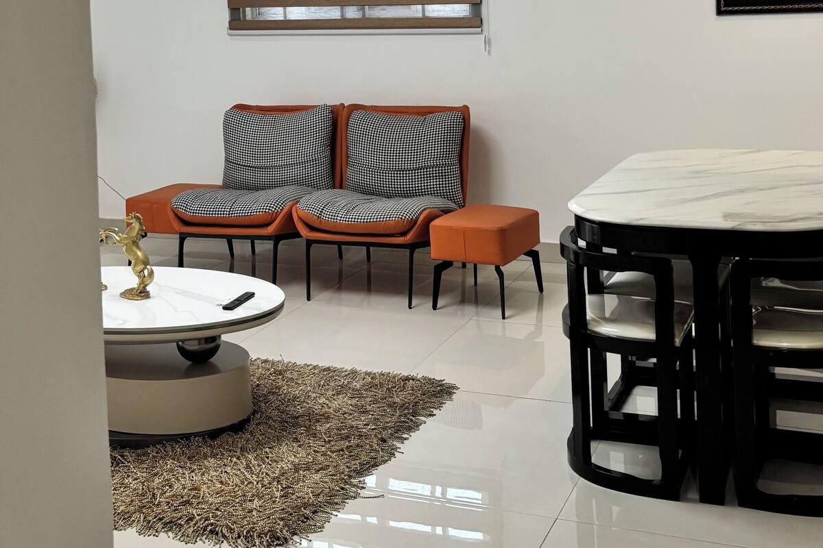 Un salon moderne avec une table basse ronde, deux fauteuils, une table à manger et une grande fenêtre avec des stores.