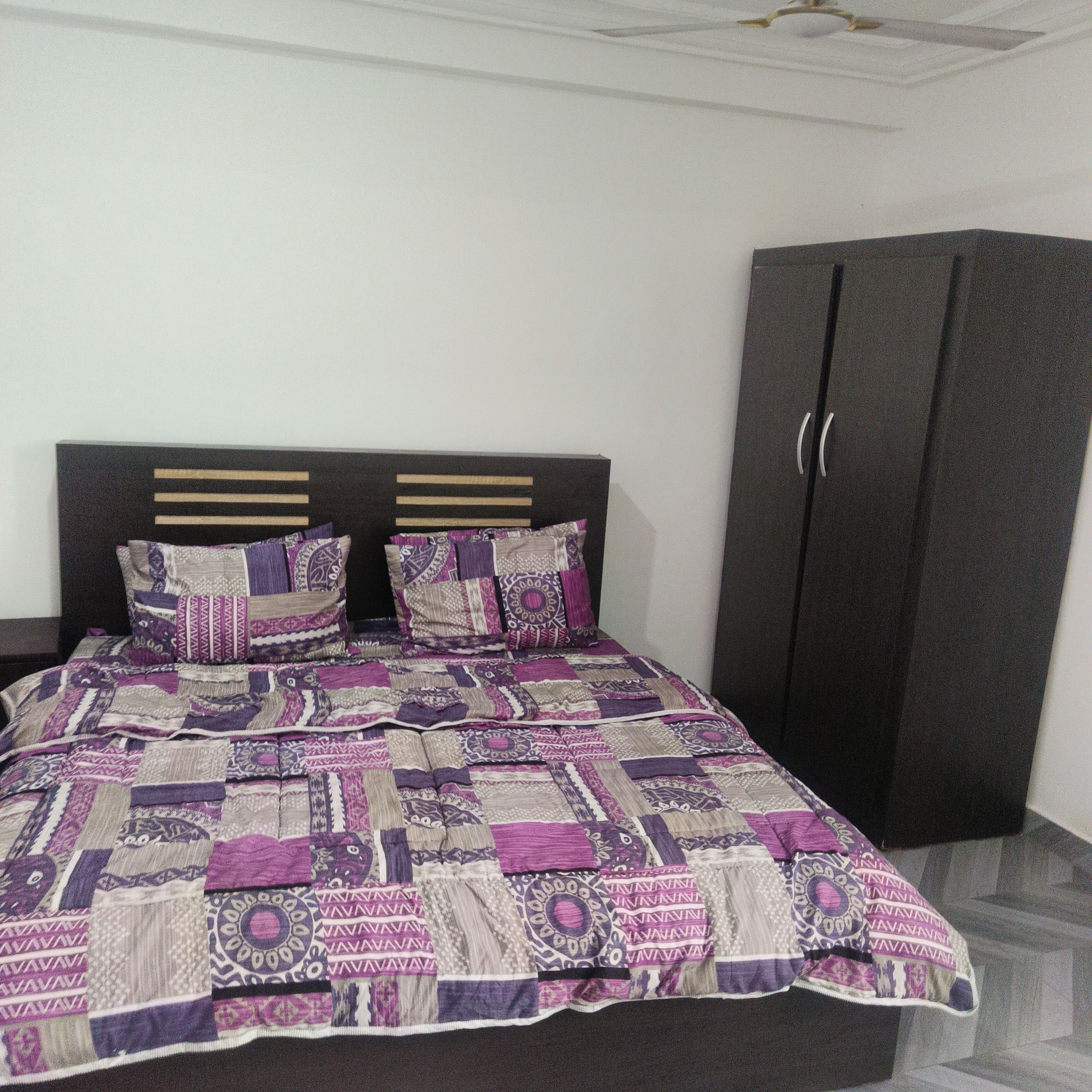 A bed with a patterned bedspread and two pillows, a dark wardrobe, and a white ceiling with a decorative element.