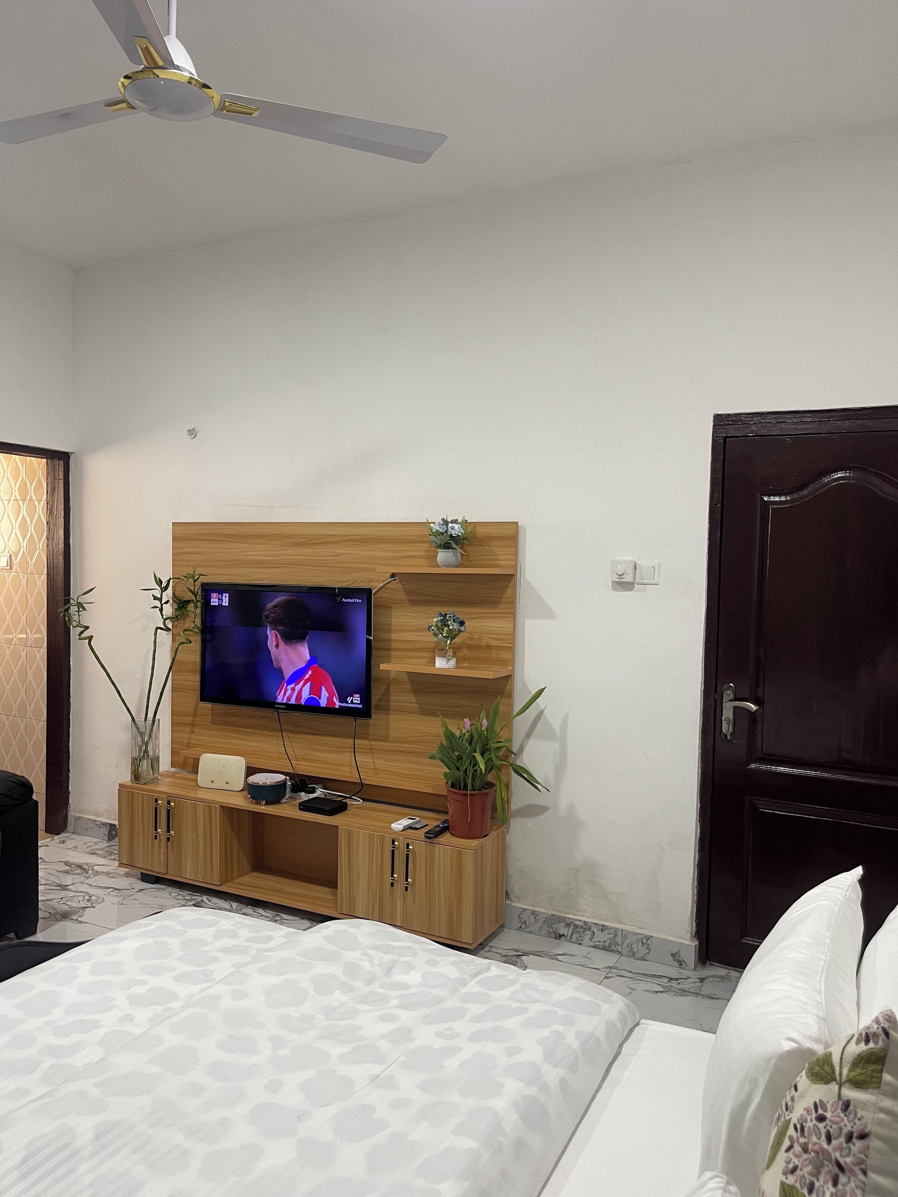 Une chambre avec un téléviseur fixé sur un meuble mural en bois, un lit avec une literie à motifs, une porte en bois foncé et un ventilateur de plafond.
