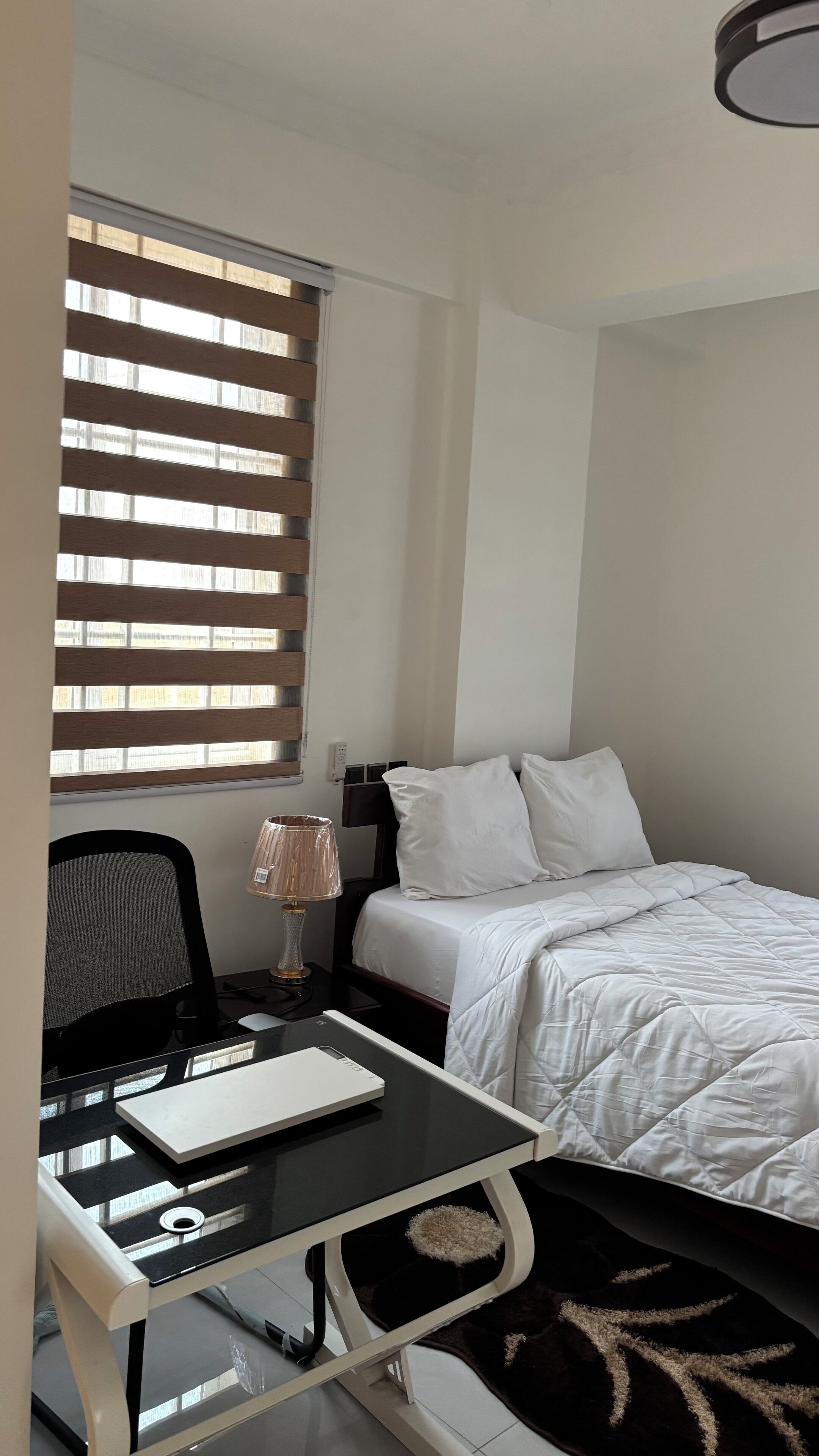 Une chambre d’hôtel avec un lit, un bureau avec un ordinateur, une chaise et une fenêtre avec des stores.