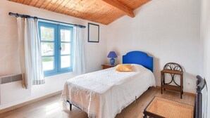 Un dormitorio con techo de madera, una cama con cabecera azul, una mesita de noche con lámpara y una silla de mimbre.