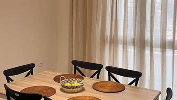 Zona pranzo con tavolo in legno, sedie nere, sottopiatti intrecciati e un cesto di frutta.