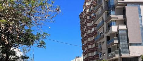 A street with parked cars, a multi-story building with balconies, and a clear blue sky.