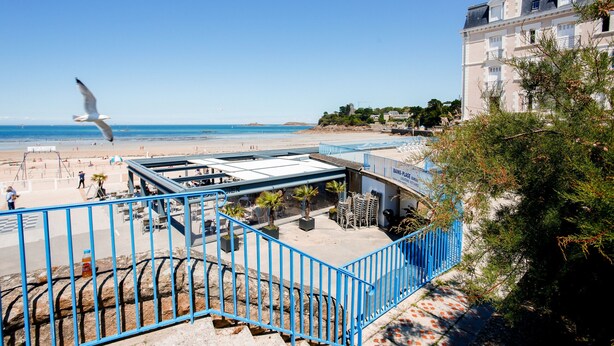 Ein Hotel an der Küste mit Blick auf den Strand, einem blauen Geländer und wolkenlosem Himmel.