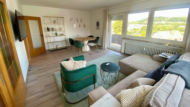 Ein geräumiger Wohnbereich mit einer Couch, einem Esstisch mit Stühlen, einem an der Wand montierten Fernseher und einem großen Fenster mit Blick auf Grünfläche.