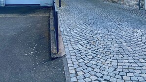 Eine Kopfsteinpflasterstraße führt zu einer Kirche, begleitet von einer Steinmauer auf der einen Seite und einem weißen Gebäude mit Garage auf der anderen.