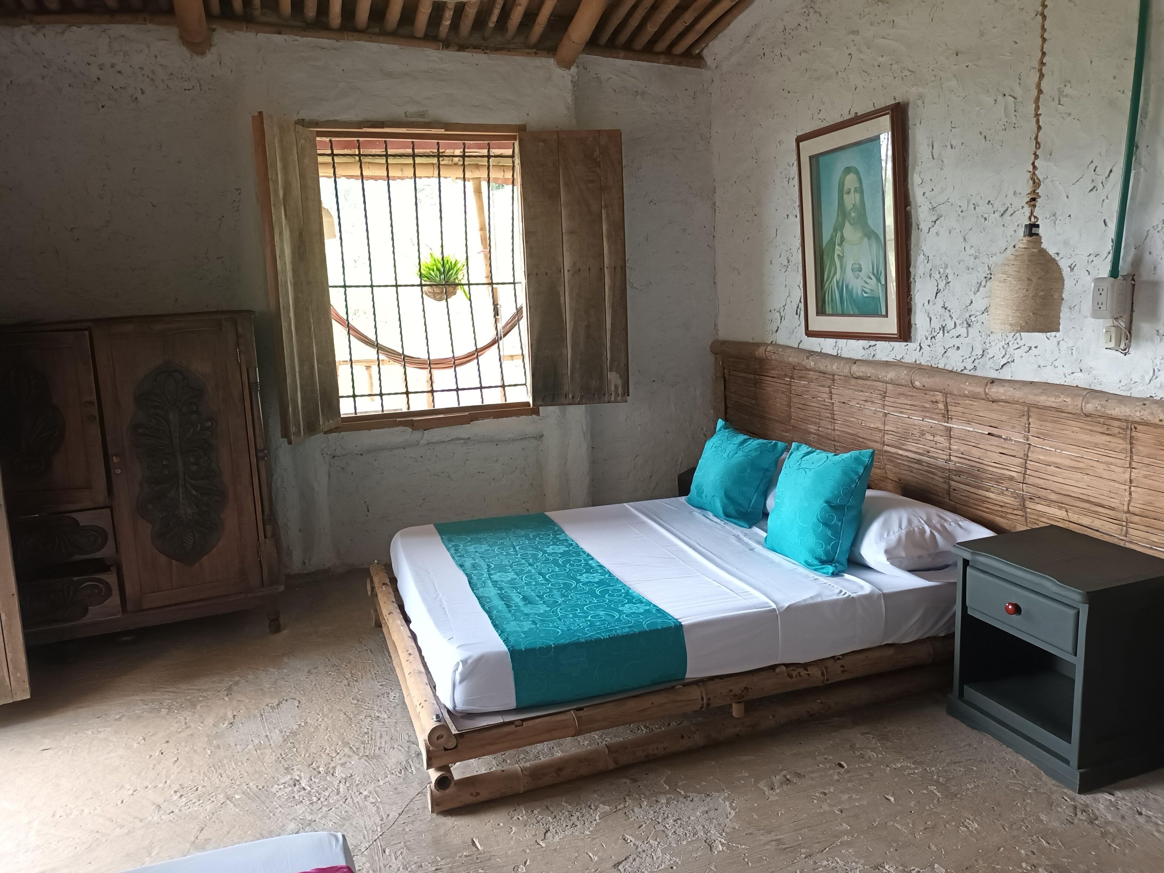 A rustic room with a wooden bed, a small bedside table, a wardrobe, and a window with bars.
