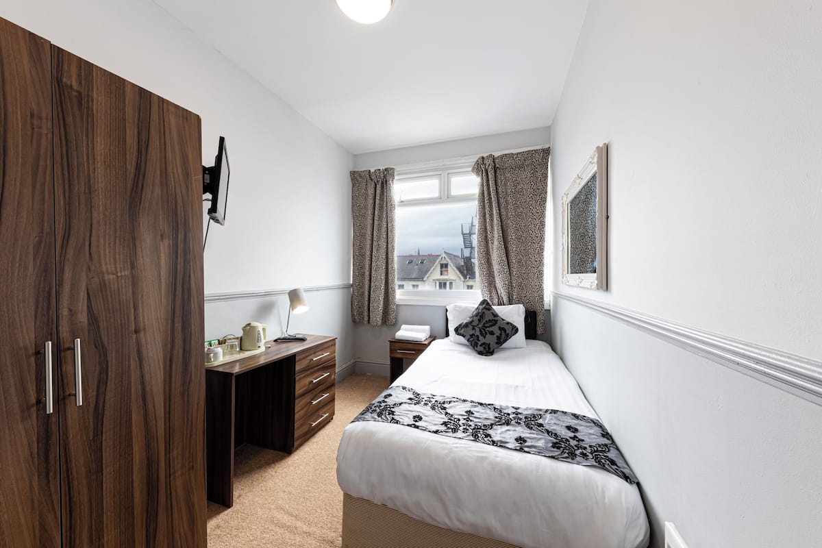 Quarto com guarda-roupa de madeira, escrivaninha com luminária, cama com colcha estampada e janela com cortinas.