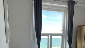 Ein Schlafzimmer mit Bett, Nachttisch, Lampe, einem Fenster mit Vorhängen und Blick auf das Meer.