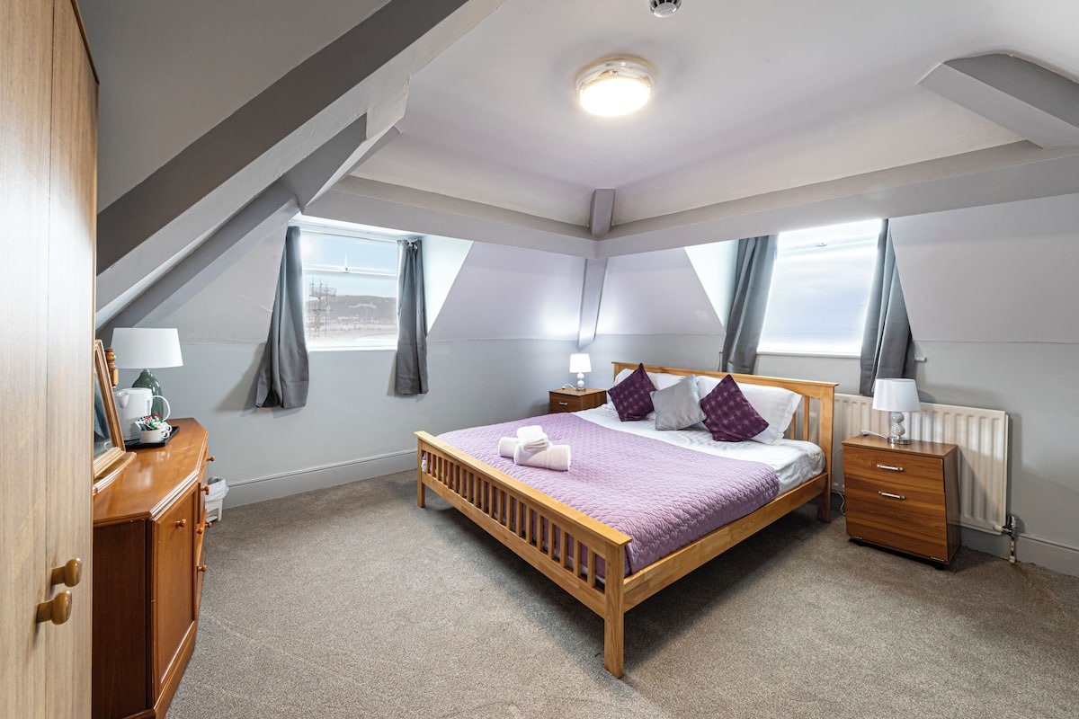 Quarto com cama de madeira, mesa de cabeceira, cômoda e janela com cortinas.