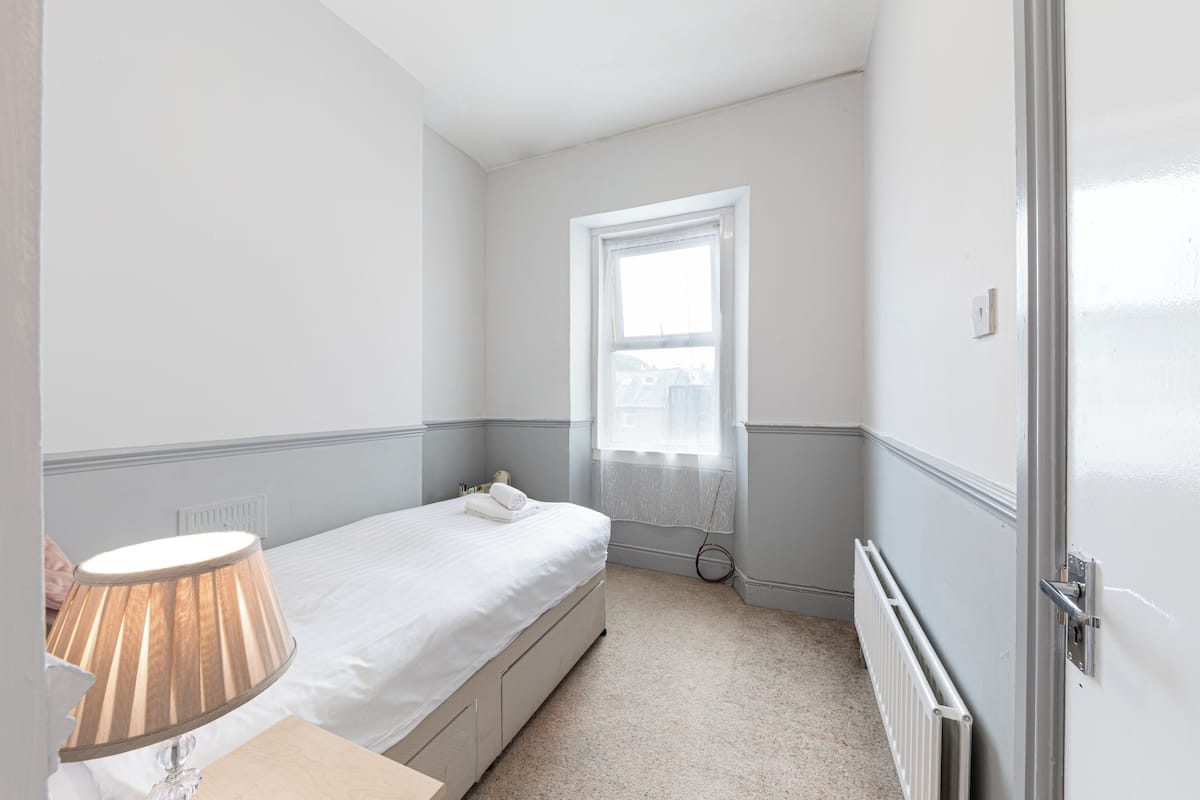 Um quarto pequeno e bem arrumado com uma cama de solteiro, mesa de cabeceira, abajur, radiador e uma janela com vista para edifícios.