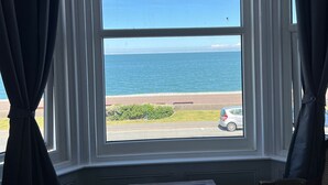 Ein Zimmer mit einem großen Erkerfenster mit Blick auf Strand und Meer. Im Vordergrund befinden sich ein kleiner Tisch und zwei Stühle.