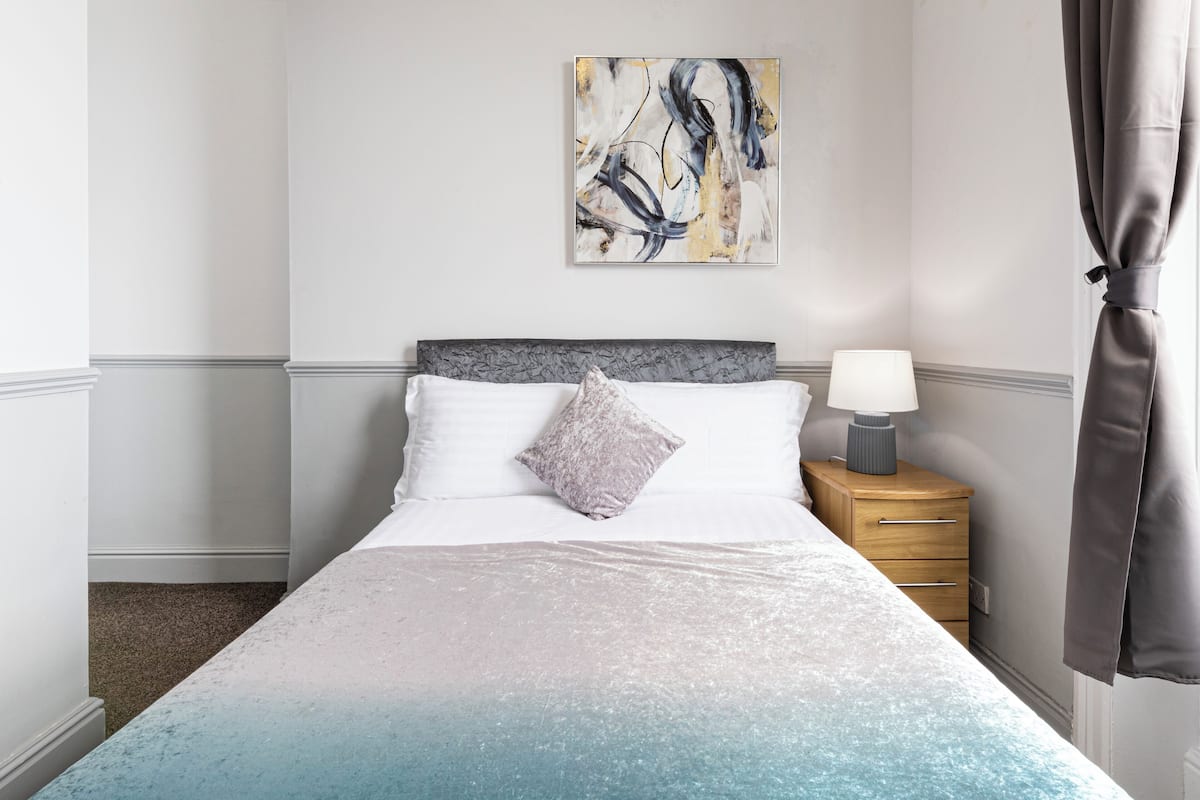 Uma cama bem arrumada com um cobertor degradê, um travesseiro, uma mesa de cabeceira com abajur e um quadro na parede.