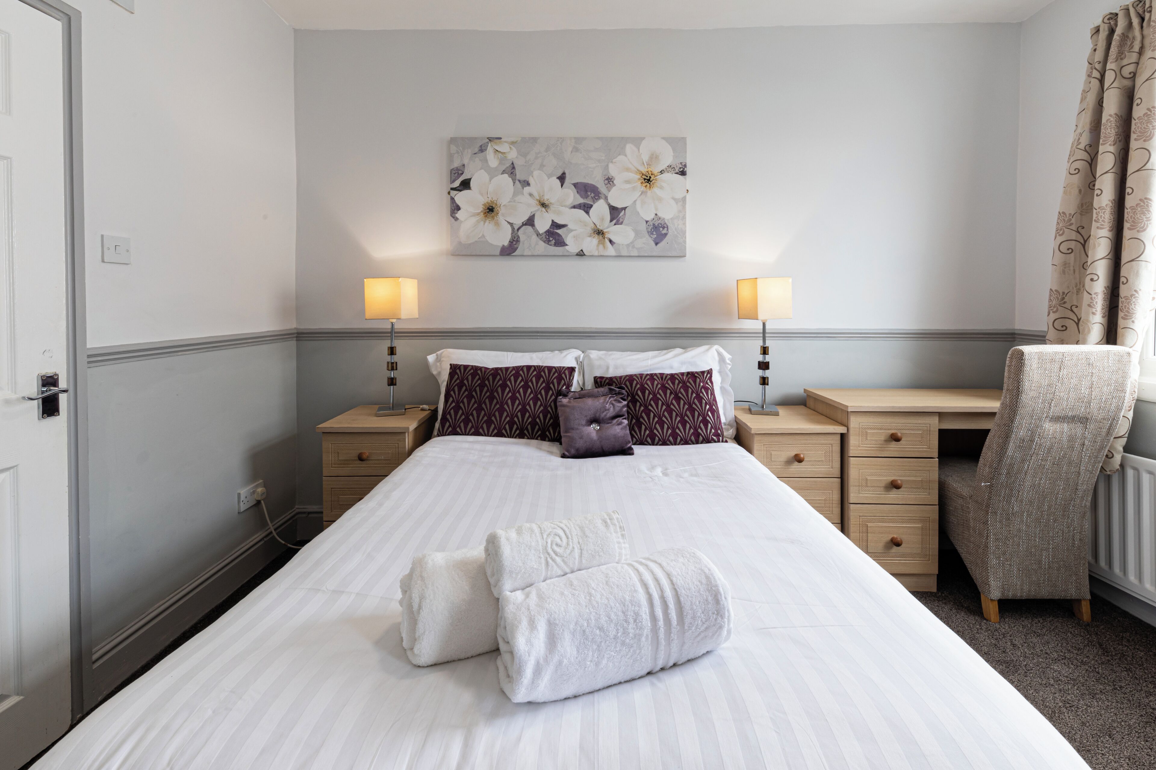 A neatly made bed with white linens and two rolled towels, a desk with a chair, and a wall-mounted lamp.