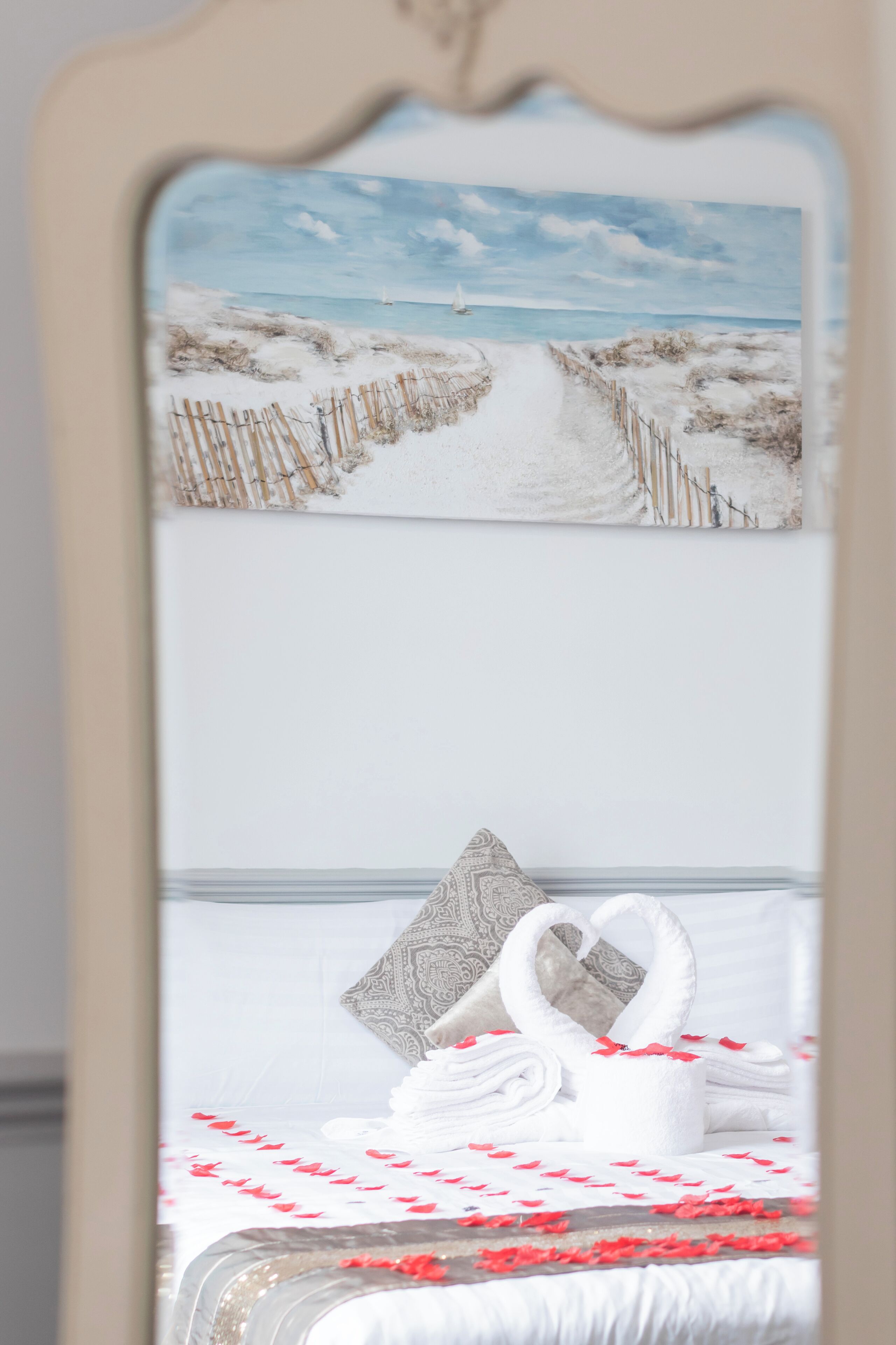 A bed with a swan-shaped towel and red rose petals, a patterned pillow, and a painting of a beach path.