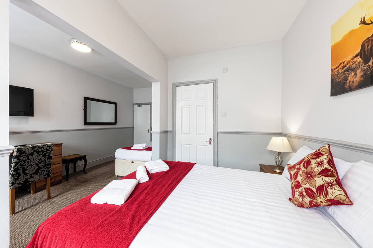 Quarto de hotel com cama, penteadeira, cadeira, espelho, televisão e porta.