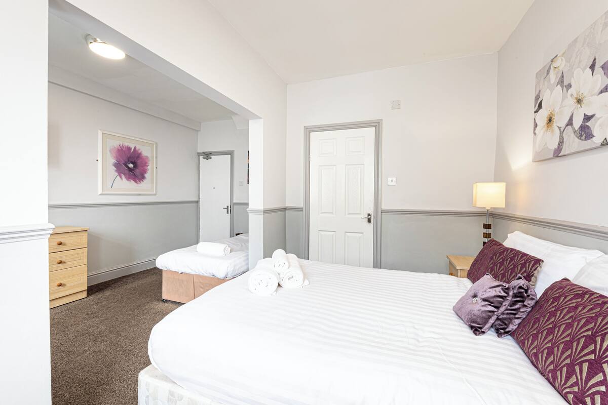 Quarto de hotel com uma cama grande, mesa de cabeceira, cómoda e porta.