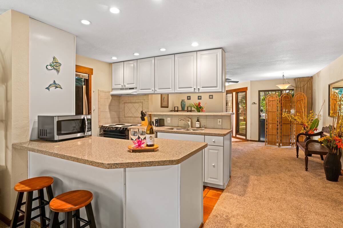 A spacious kitchen with white cabinets, a large island with stools, a microwave, and a wine bottle.