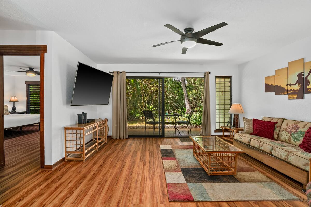 A spacious living room with a ceiling fan, a flat-screen TV, a sofa, a coffee table, and a view of a garden through glass doors.